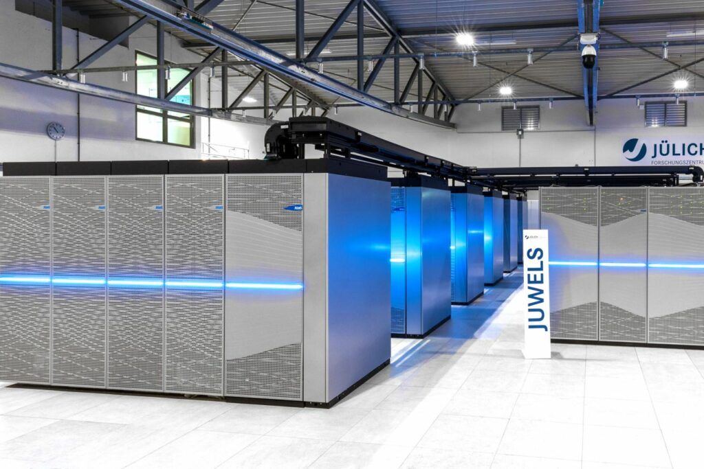 Jewls Supercomputer im JSC. Hauptsächlich silberner Supercomputer mit hellen blauen Lichtern an der Vorderseite. Stehend im offenen Raum.